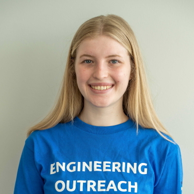 A smiling young woman wearing a blue t-shirt with the text "ENGINEERING OUTREACH."