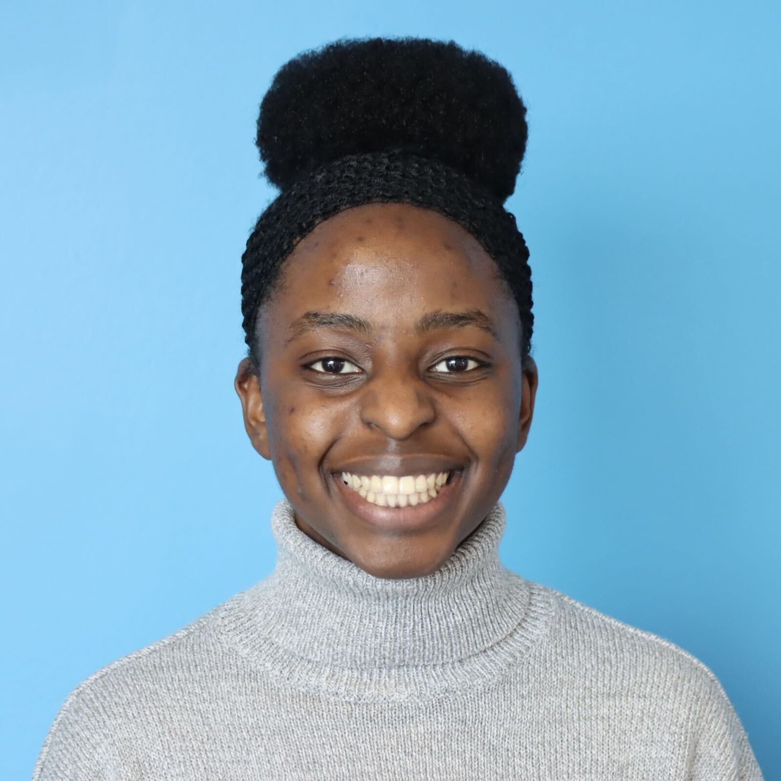 Smiling person with hair in a high bun, wearing a gray turtleneck sweater against a light blue background.