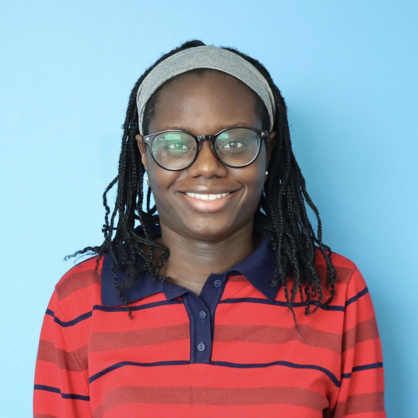 Smiling person with glasses and a gray headband, wearing a red-and-navy striped shirt against a light blue background.