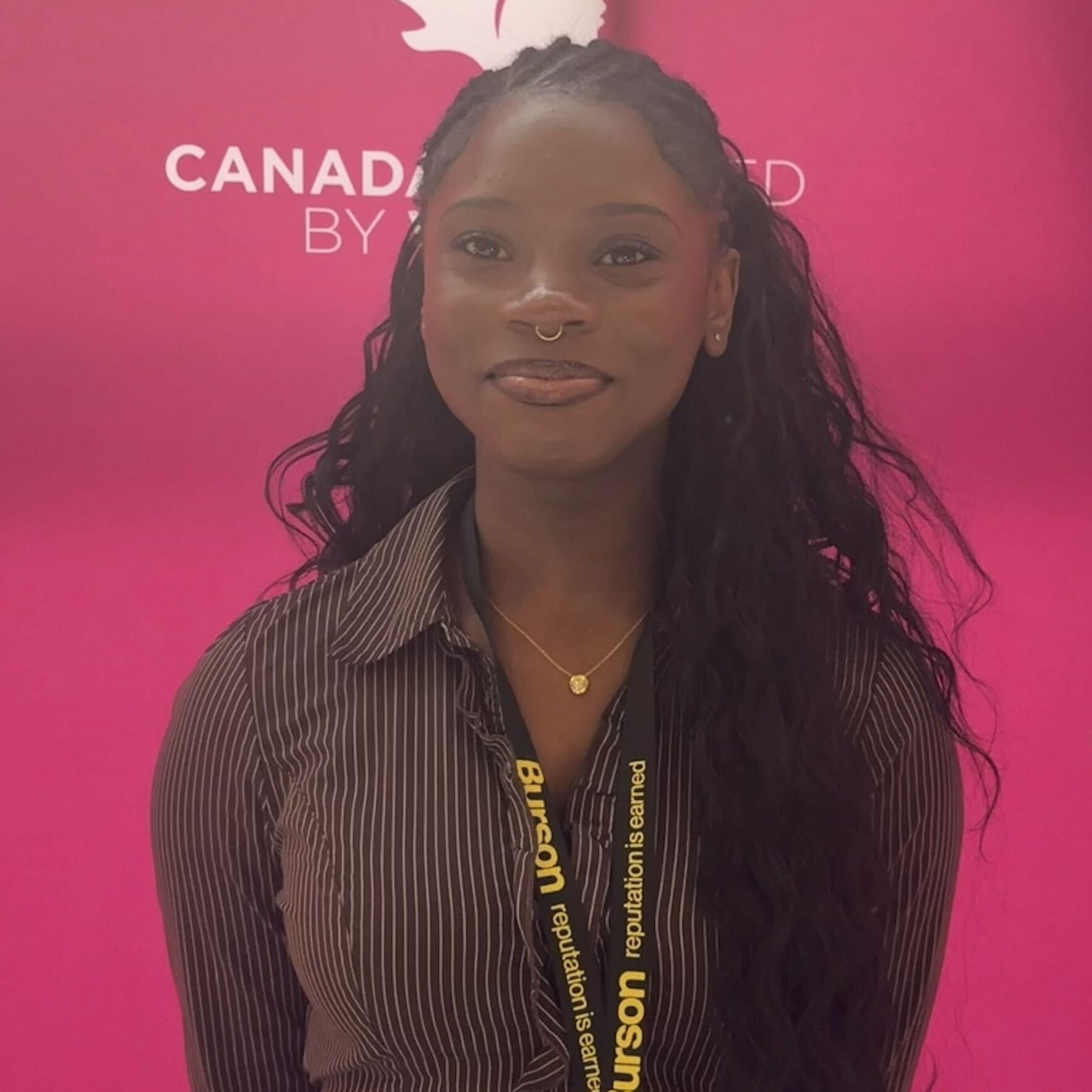 Portrait of a woman wearing a striped shirt and a lanyard, standing in front of a bright pink backdrop with partial "CANADA" text.