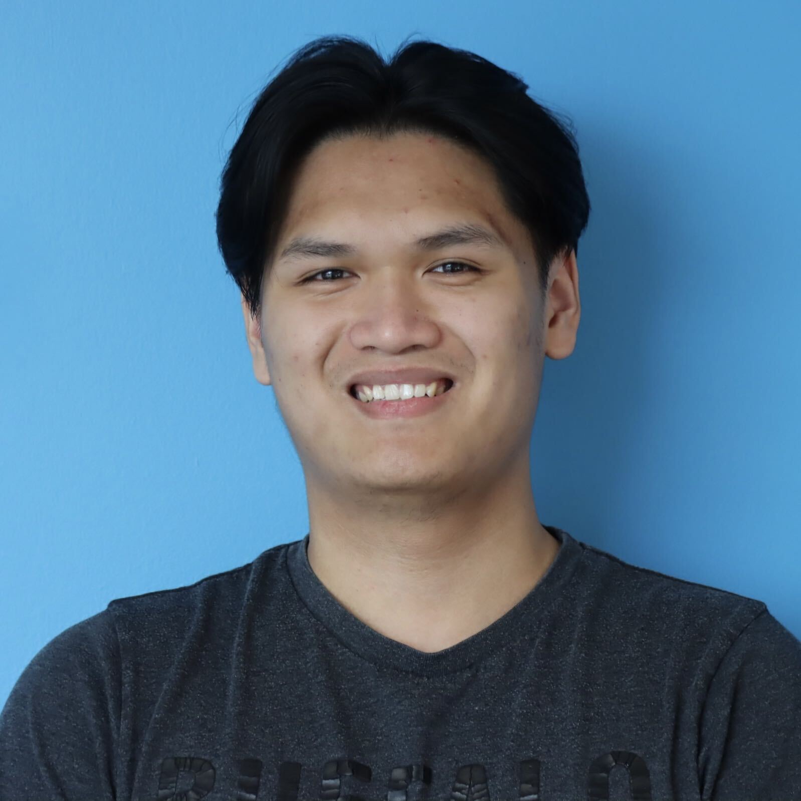 Smiling person with dark hair wearing a dark gray T-shirt against a solid blue background.