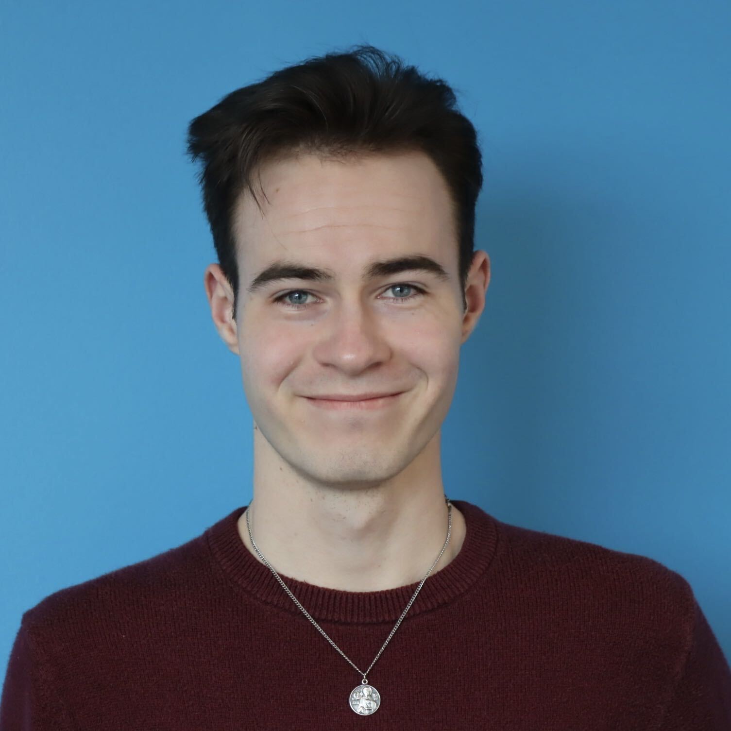 Head-and-shoulders portrait of a smiling young man with dark hair against a blue background, wearing a maroon sweater and a necklace with a round pendant.