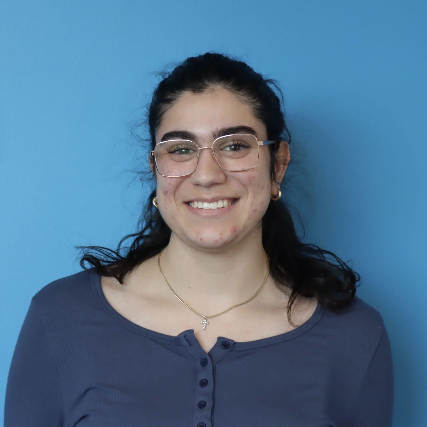 Smiling person wearing glasses and a necklace in a head-and-shoulders portrait against a bright blue background.