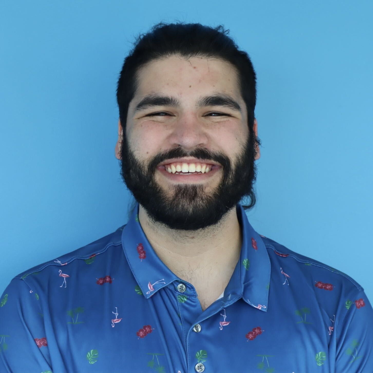 Smiling bearded person in a blue patterned shirt against a solid light-blue background.