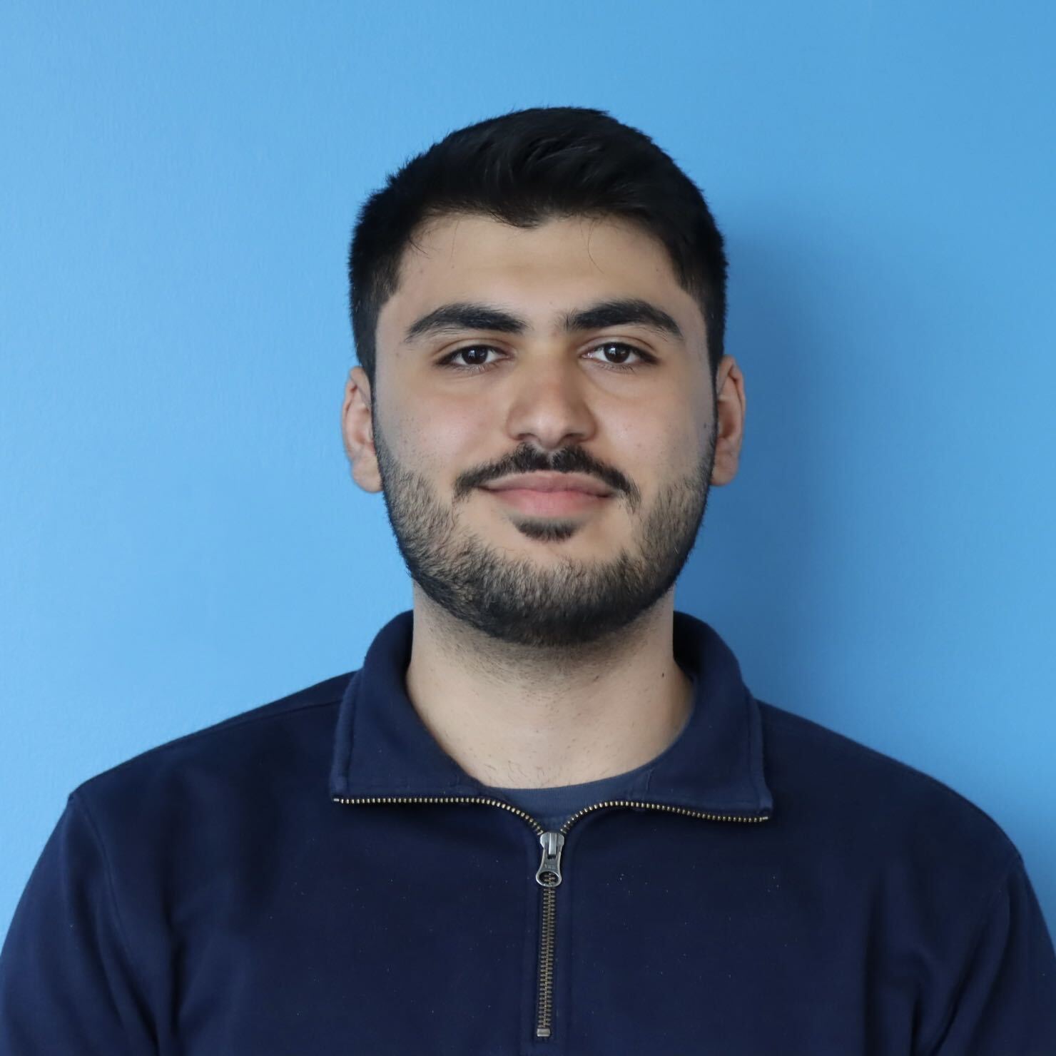Head-and-shoulders portrait of a man with short dark hair and a trimmed beard, wearing a navy zip-up top against a bright blue background.