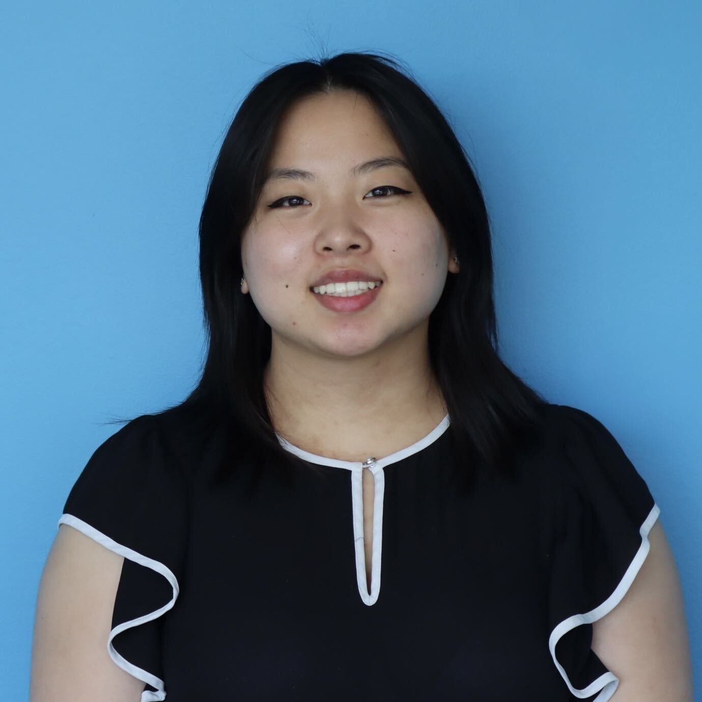 Smiling person with shoulder-length dark hair wearing a black top with white trim, posed against a blue background.