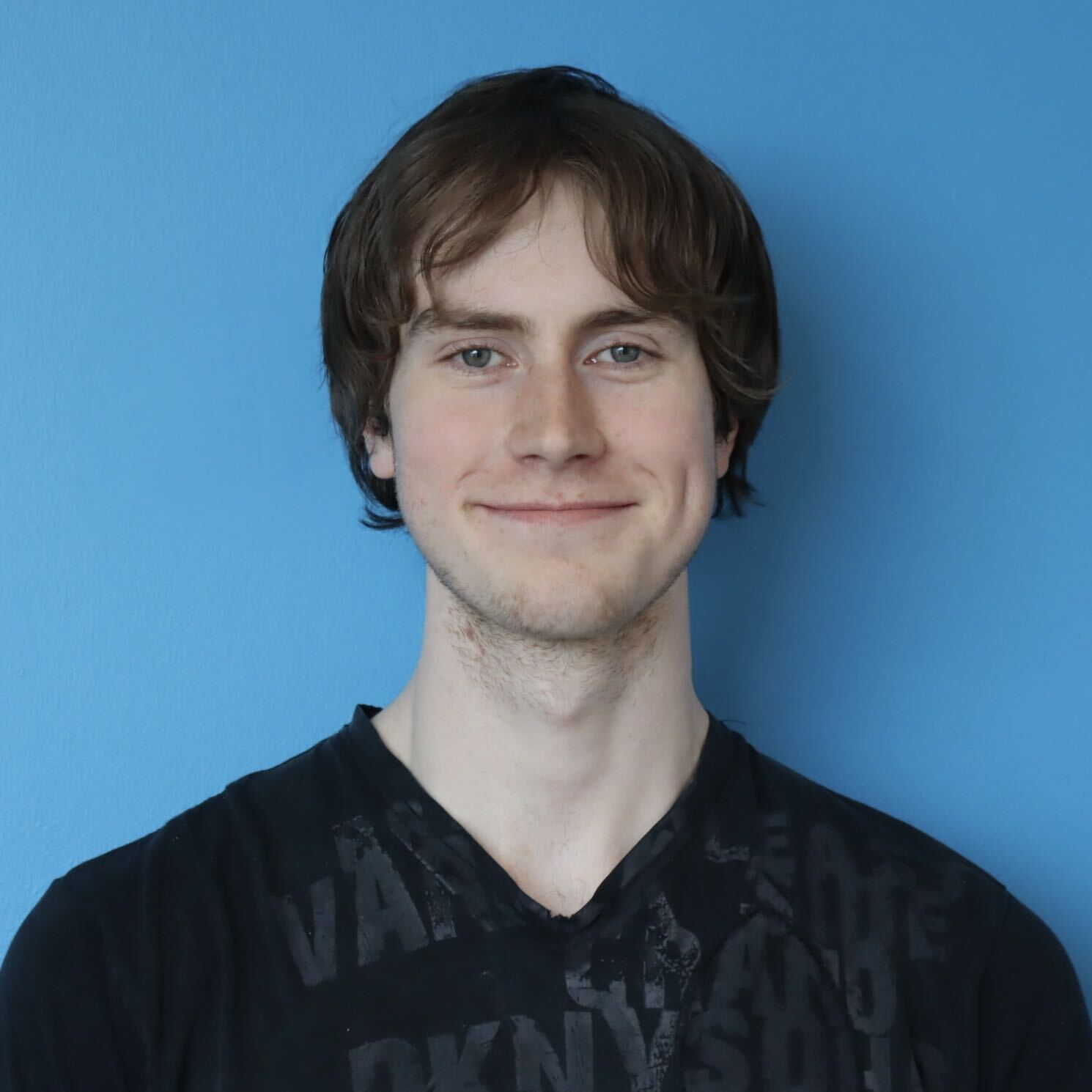 Head-and-shoulders portrait of a young man with medium brown hair, wearing a black T-shirt, against a bright blue background.