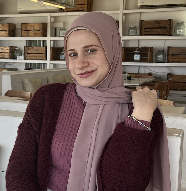 A young woman wearing a pink hijab and a maroon sweater poses confidently, smiling at the camera in a cozy café setting.