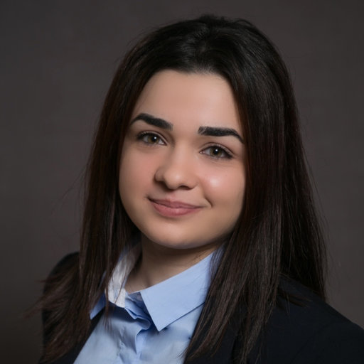 Professional headshot of a young woman with long dark hair, wearing a blue shirt and a black blazer, smiling softly.