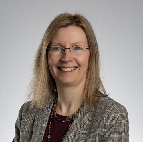 Professional headshot of a woman with long, straight blonde hair, wearing glasses and a plaid blazer, smiling at the camera.