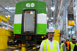 Construction worker in a hard hat and reflective vest giving a thumbs-up inside an industrial facility, with coworkers working in the background.