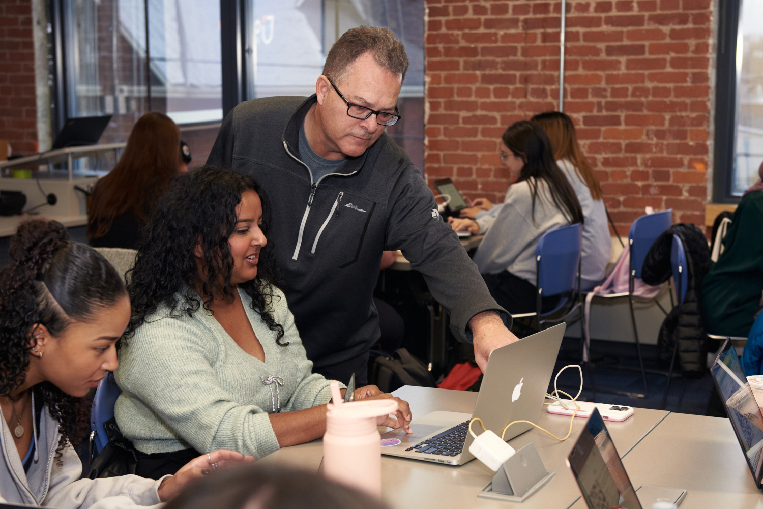 Brian Errey, a mathematics instructor in the Bachelor of Education program, assists a student with a laptop in a classroom setting, while other students work on their devices in the background.