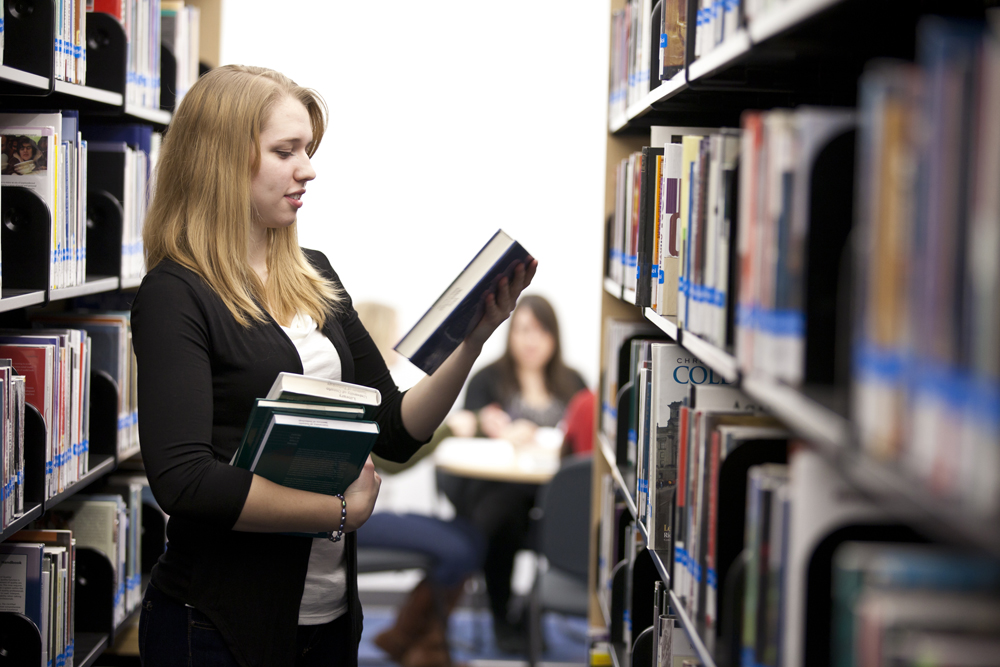 A student holding books on her right hand while reading a book on her left hand in the library aisle