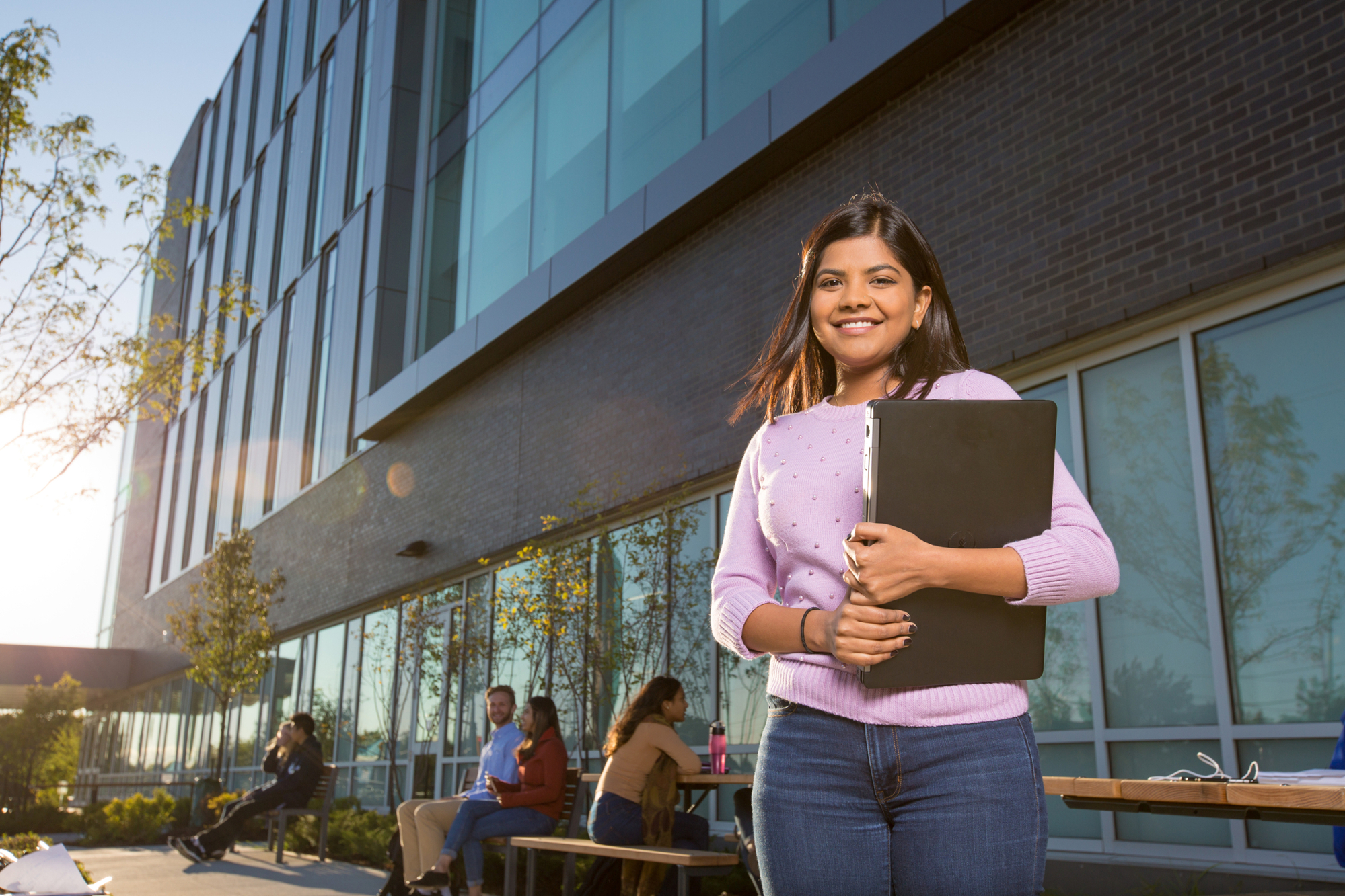 A student standing in front of the SIRC building holding a laptop. 