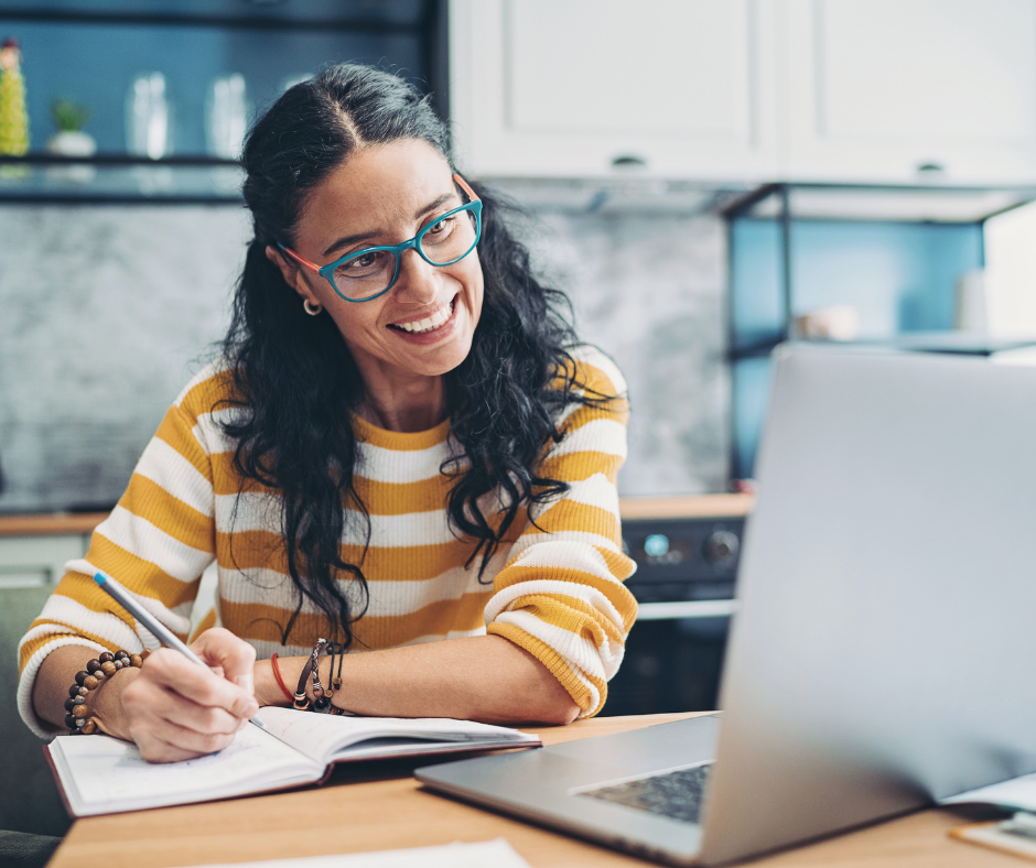 An image of a person smiling and sitting in front of a laptop taking notes. 